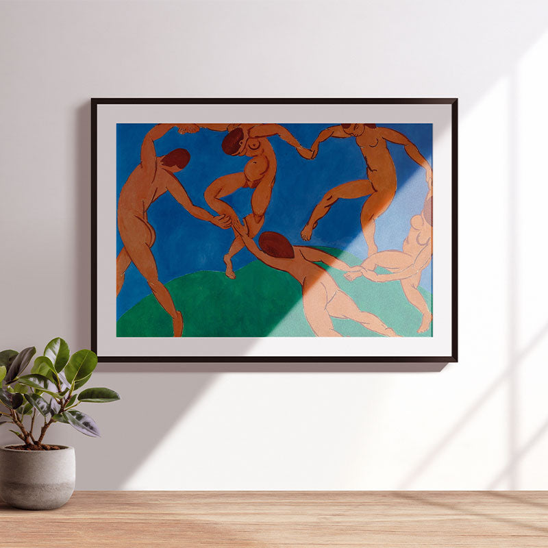 Quadro Decorativo A Dança Matisse com moldura preta e passe-partout branco, pendurado em parede clara com luz natural e planta ao lado.