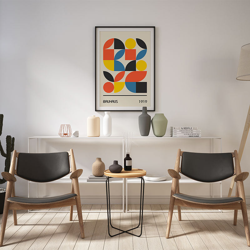 Quadro Decorativo Geométrico Bauhaus com moldura preta pendurado em parede branca, entre cadeiras de madeira e prateleira decorada.